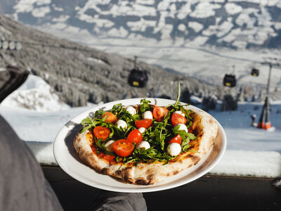 Hand hält Teller mit Pizza belegt mit Rucola, Kirschtomaten und Mozzarellakugeln vor verschneiter Berglandschaft mit Gondelbahn
