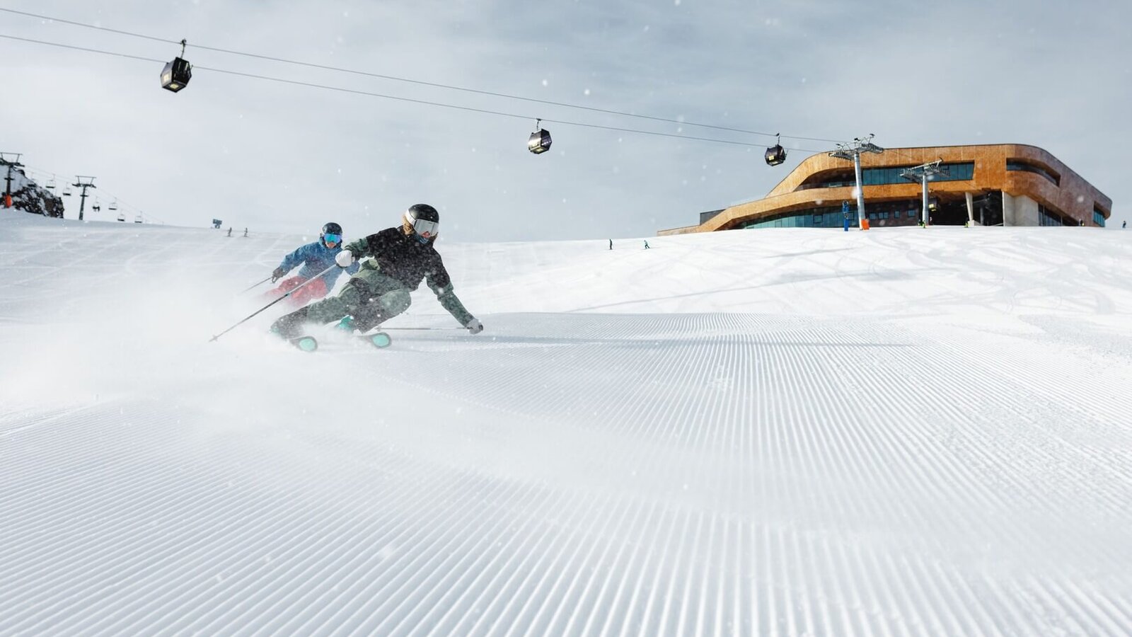 Zwei Skifahrer fahren auf einer frisch präparierten Piste mit Gondeln und einer Bergstation im Hintergrund