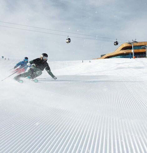 Zwei Skifahrer fahren auf einer frisch präparierten Piste mit Gondeln und einer Bergstation im Hintergrund