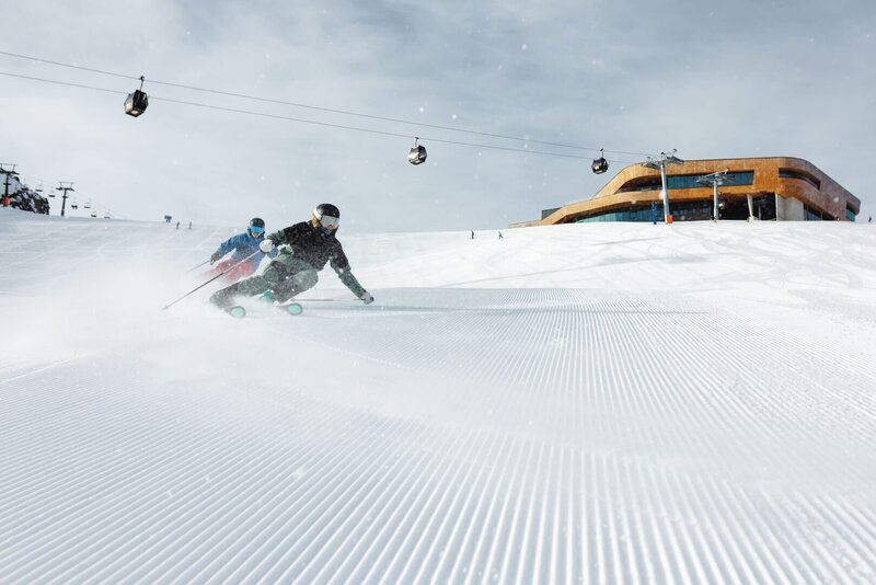 Zwei Skifahrer fahren auf einer frisch präparierten Piste mit Gondeln und einer Bergstation im Hintergrund