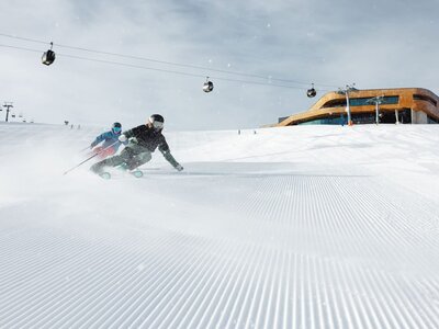 Zwei Skifahrer fahren auf einer frisch präparierten Piste mit Gondeln und einer Bergstation im Hintergrund