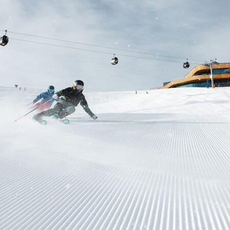 Zwei Skifahrer fahren auf einer frisch präparierten Piste mit Gondeln und einer Bergstation im Hintergrund