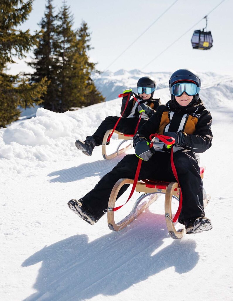 Zwei Personen mit Helm und Winterkleidung sitzen auf Schlitten auf einer verschneiten Piste, im Hintergrund Bäume und eine Gondelbahn