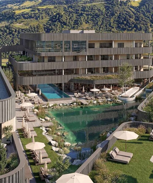 Mehrstöckiges modernes Hotelgebäude mit Holzfassade vor bewaldeten Berg und einem natürlichen Schwimmteich mit Liegestühlen und Sonnenschirmen auf einer Wiese