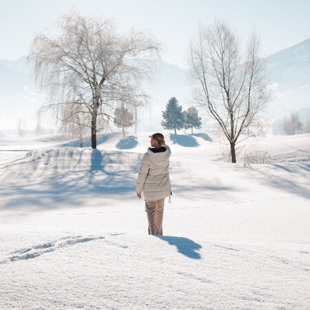 Person mit heller Winterjacke steht auf verschneiter Fläche vor kahlen Bäumen und Bergen im Hintergrund