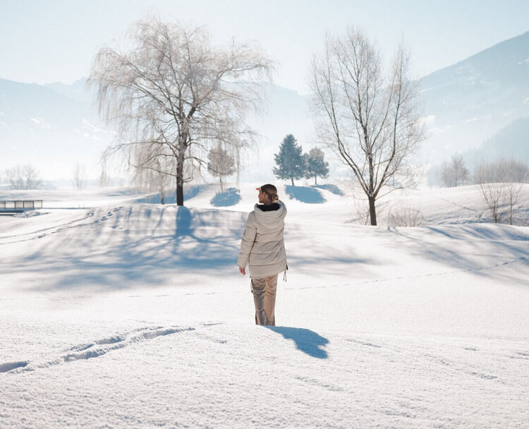Person mit heller Winterjacke steht auf verschneiter Fläche vor kahlen Bäumen und Bergen im Hintergrund