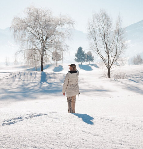 Person mit heller Winterjacke steht auf verschneiter Fläche vor kahlen Bäumen und Bergen im Hintergrund