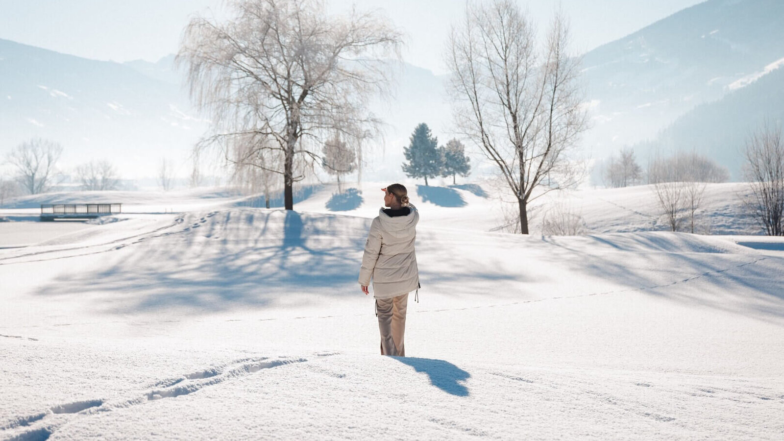 Person mit heller Winterjacke steht auf verschneiter Fläche vor kahlen Bäumen und Bergen im Hintergrund