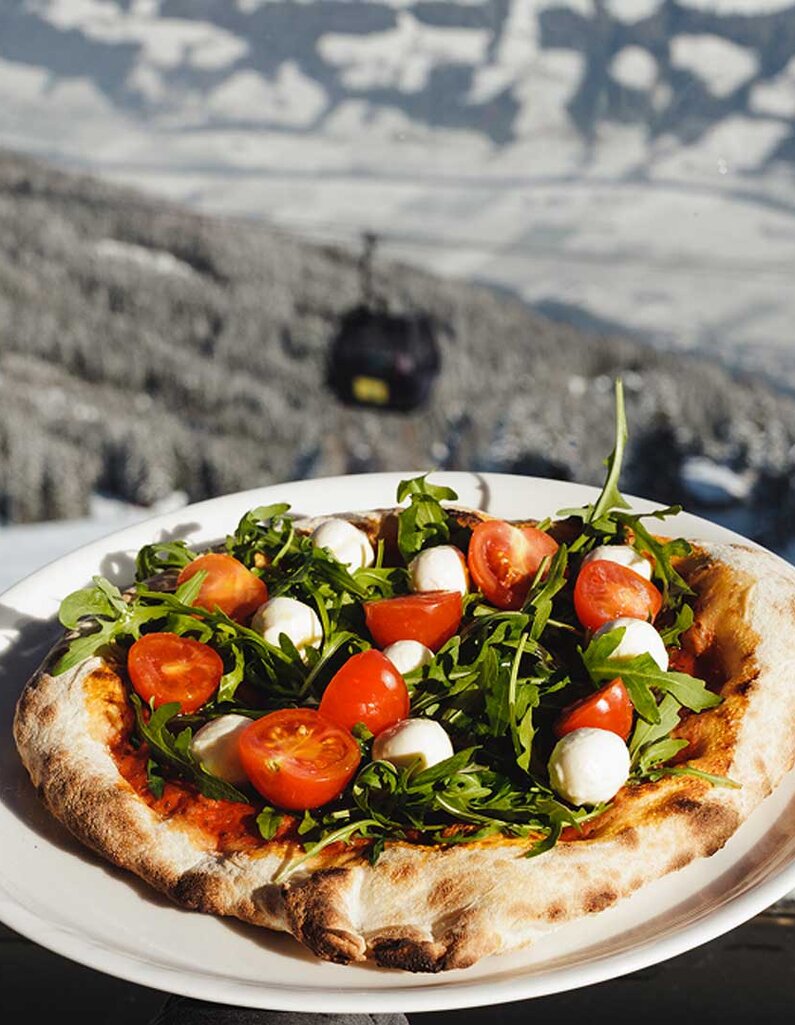 Hand hält Teller mit Pizza belegt mit Rucola, Kirschtomaten und Mozzarella vor verschneiter Berglandschaft mit Gondelbahn