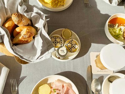 Tisch mit gedecktem Frühstück, darunter ein Brotkorb mit Brötchen, Teller mit Aufschnitt und Butter, Schalen mit Müsli und Honig, Besteck und Servietten.