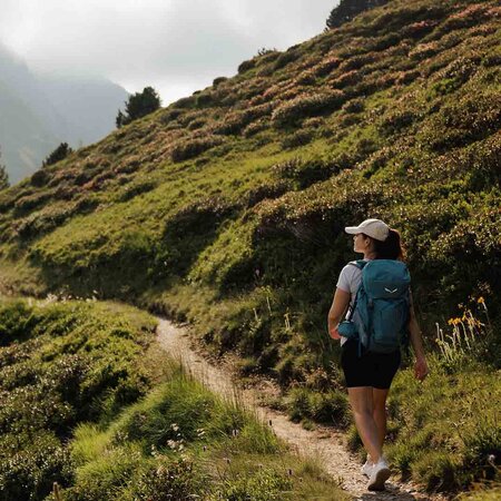Wanderin mit Rucksack und Cap auf schmalem Pfad am bewachsenen Berghang mit Bäumen und Nebel im Hintergrund