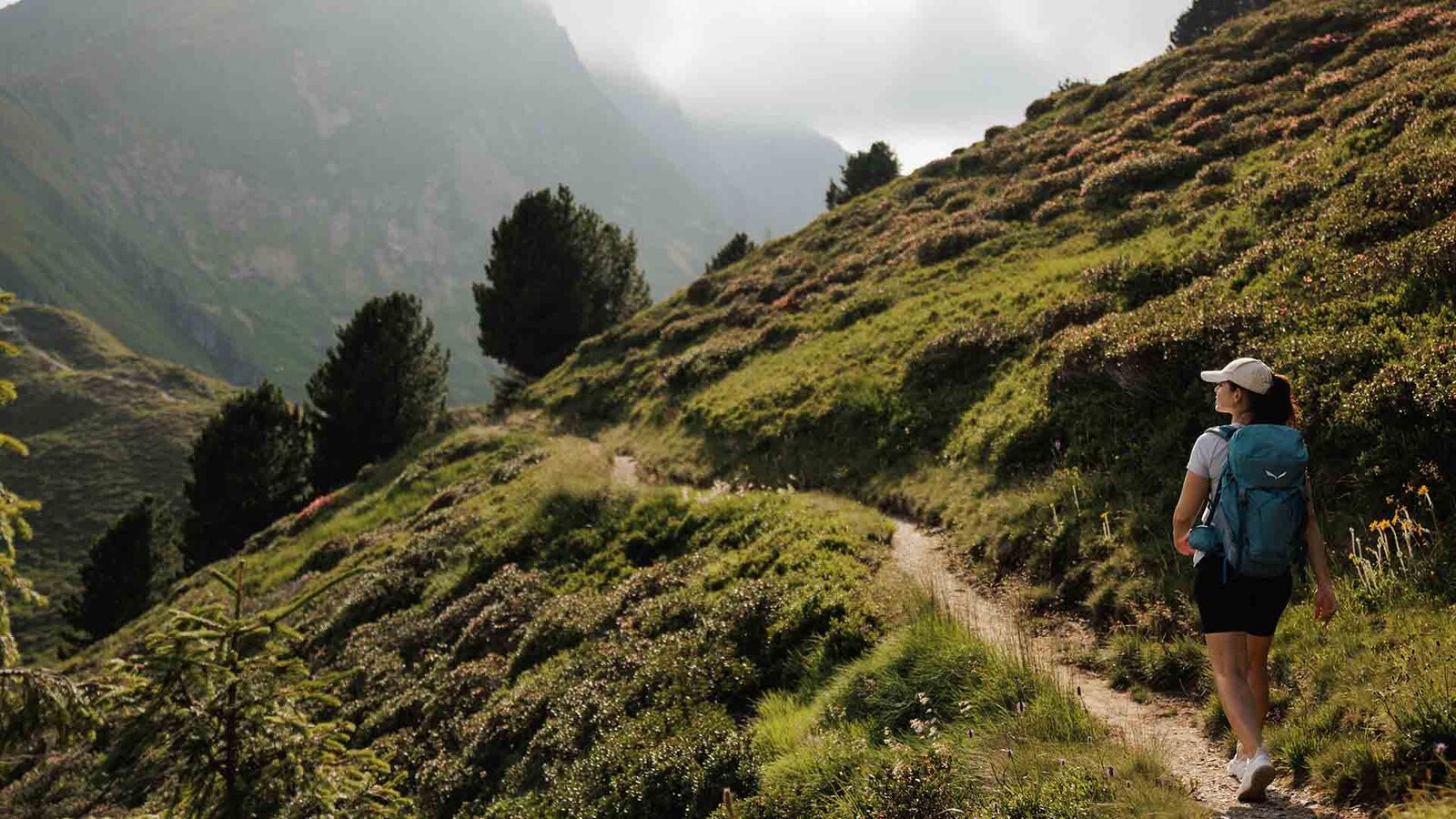 Wanderin mit Rucksack und Cap auf schmalem Pfad am bewachsenen Berghang mit Bäumen und Nebel im Hintergrund