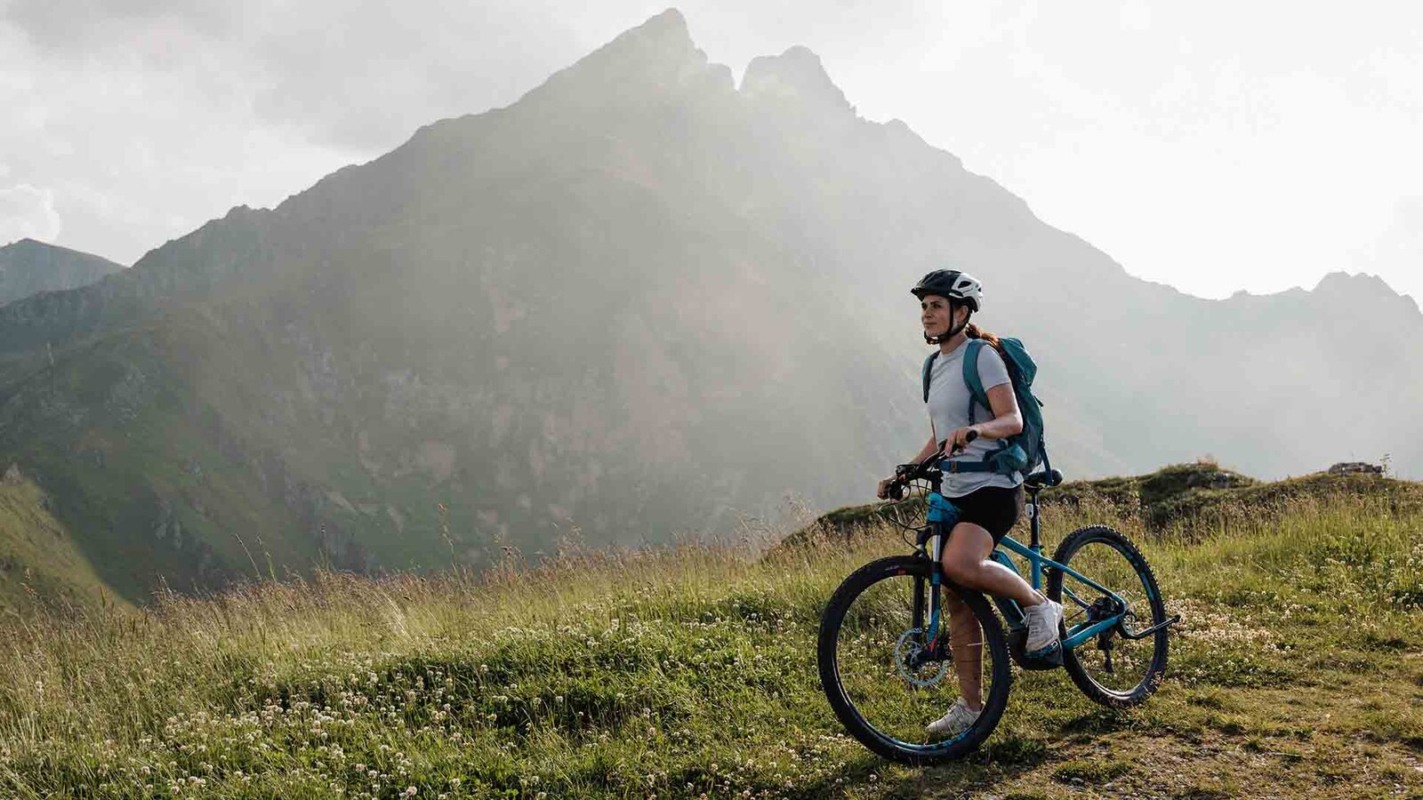 Fahrradfahrerin mit Helm und Rucksack steht mit Mountainbike auf grasbewachsenem Hügel vor nebligen Bergen