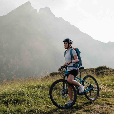 Fahrradfahrerin mit Helm und Rucksack steht mit Mountainbike auf grasbewachsenem Hügel vor nebligen Bergen