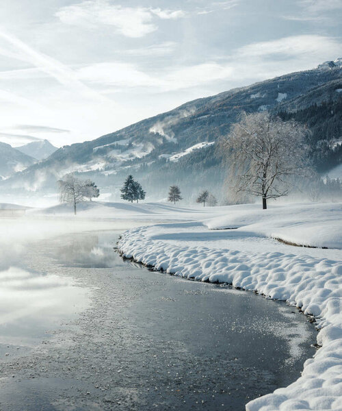Verschneite Landschaft mit zugefrorenem Fluss, einzelnen Bäumen und Bergen im Hintergrund unter bewölktem Himmel