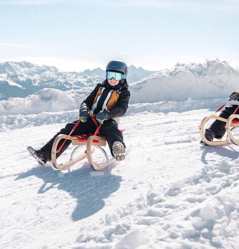 Zwei Kinder mit Helmen und Skibrillen sitzen auf Holzschlitten auf einer verschneiten Piste vor Bergpanorama