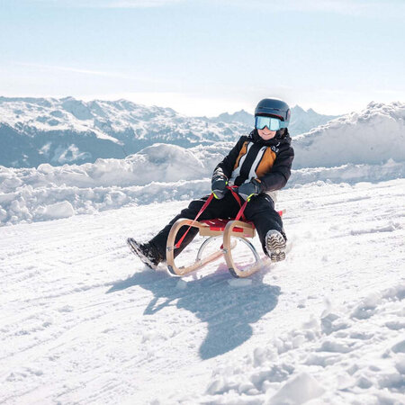 Zwei Kinder mit Helmen und Skibrillen sitzen auf Holzschlitten auf einer verschneiten Piste vor Bergpanorama