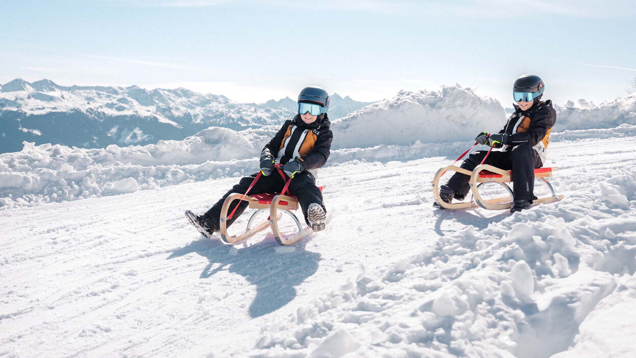 Zwei Kinder mit Helmen und Skibrillen sitzen auf Holzschlitten auf einer verschneiten Piste vor Bergpanorama
