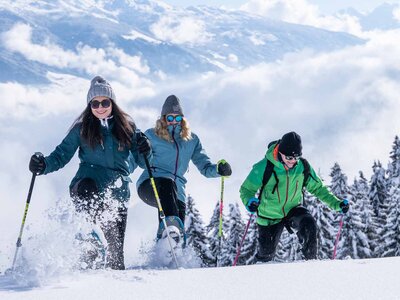 Drei Personen mit Winterkleidung und Skistöcken laufen durch tiefen Schnee vor verschneiten Tannenbäumen unter blauem Himmel mit Wolken