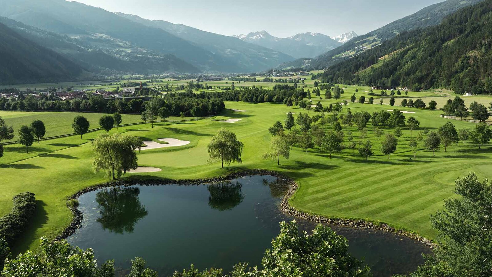 Landschaft mit Golfplatz, Teich, Bäumen und Bergen im Hintergrund